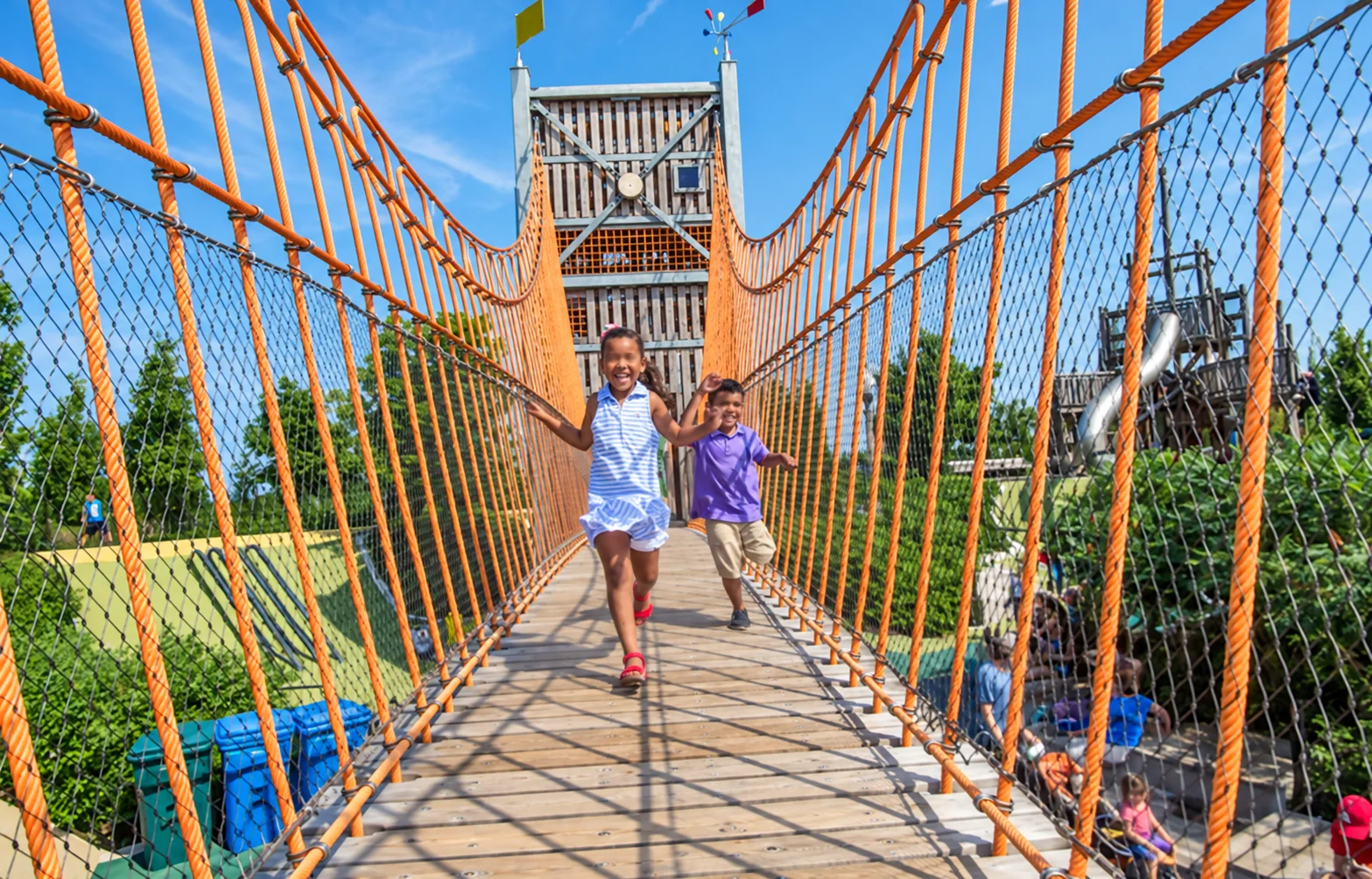 Children crossing a wooden playground bridge with orange rope sides
